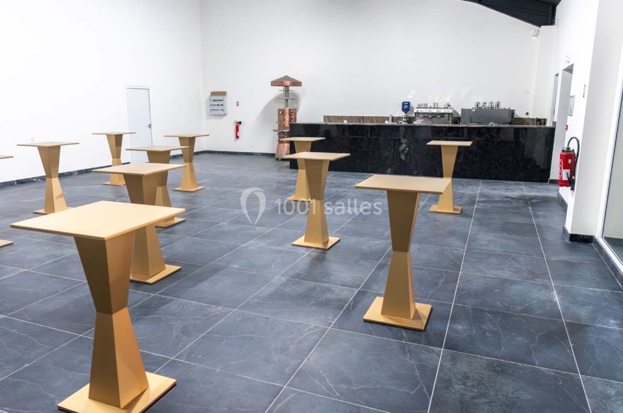 Salle vide avec sol en carrelage foncé, tables hautes dorées espacées et comptoir en arrière-plan.