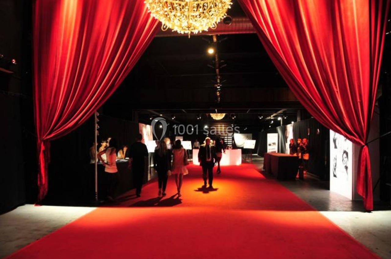 Salle sombre avec tapis rouge, rideaux rouges ouverts et lustre, accueillant des personnes lors d'un événement.