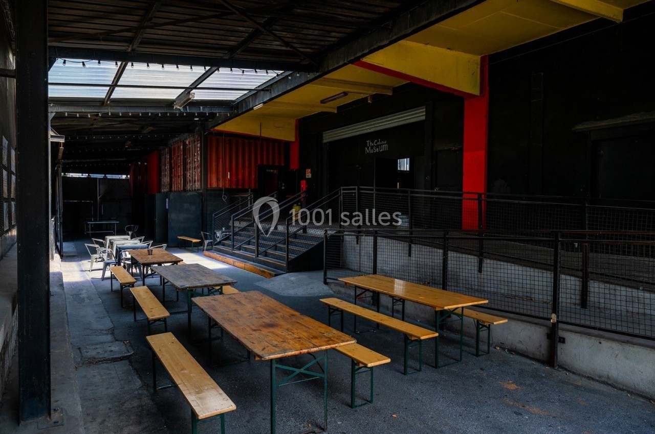 Tables et bancs en bois disposés dans une cour extérieure couverte, entourée de murs sombres et d'éléments industriels.