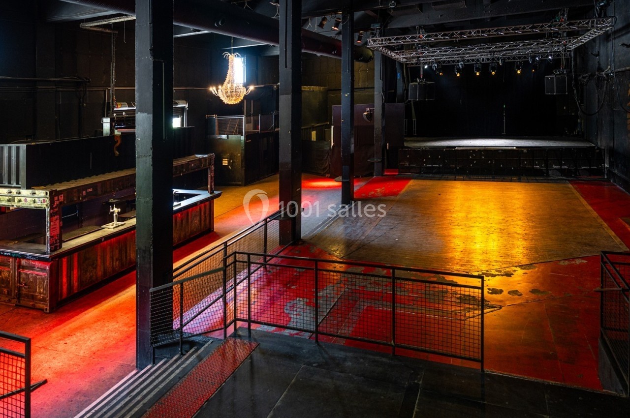 Salle de concert vide avec scène surélevée, sol rouge et noir, bar à gauche et éclairage tamisé.