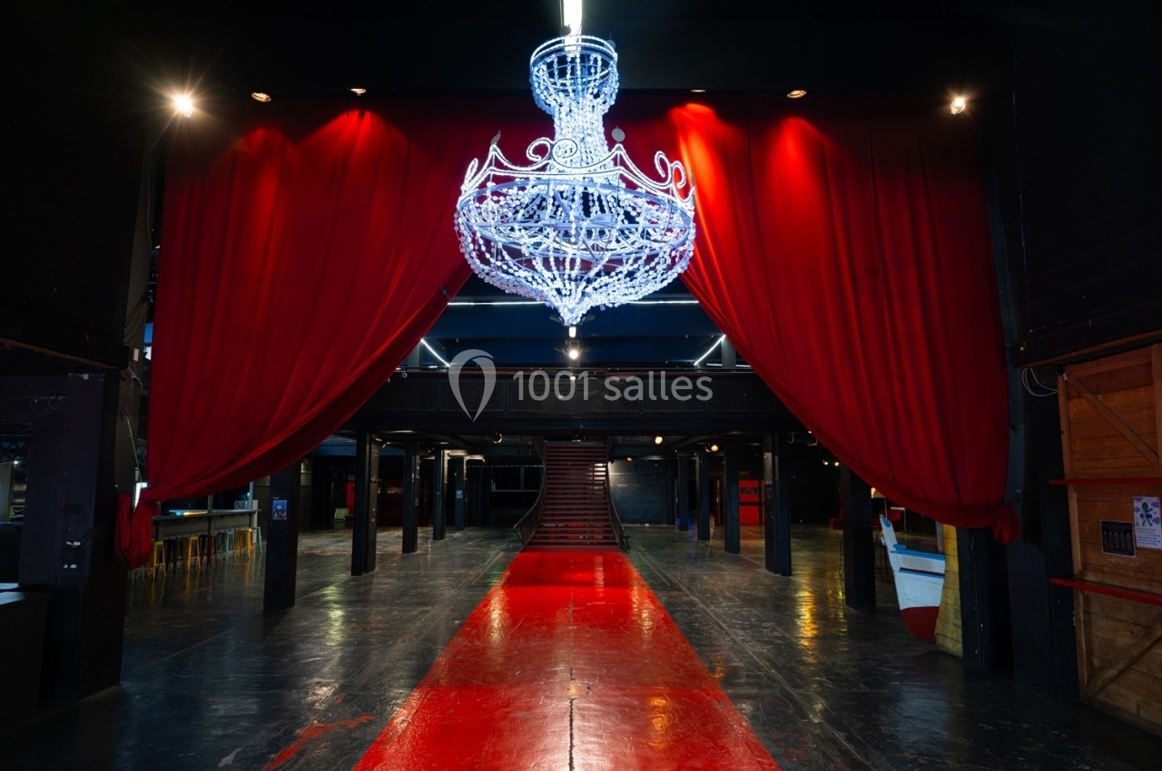 Salle sombre avec un grand lustre lumineux suspendu, rideaux rouges ouverts et tapis rouge menant à un escalier.