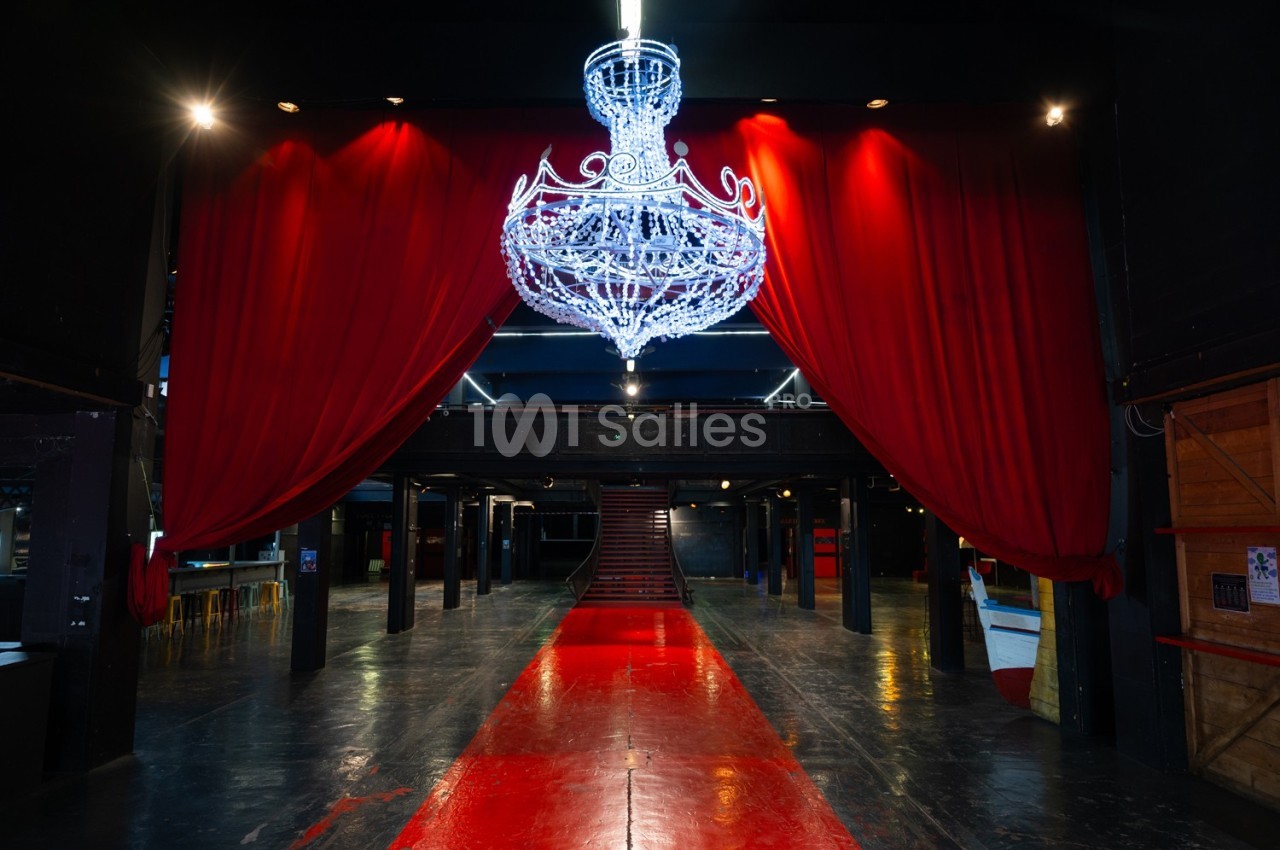 Salle sombre avec un grand lustre lumineux suspendu, rideaux rouges ouverts et tapis rouge menant à un escalier.