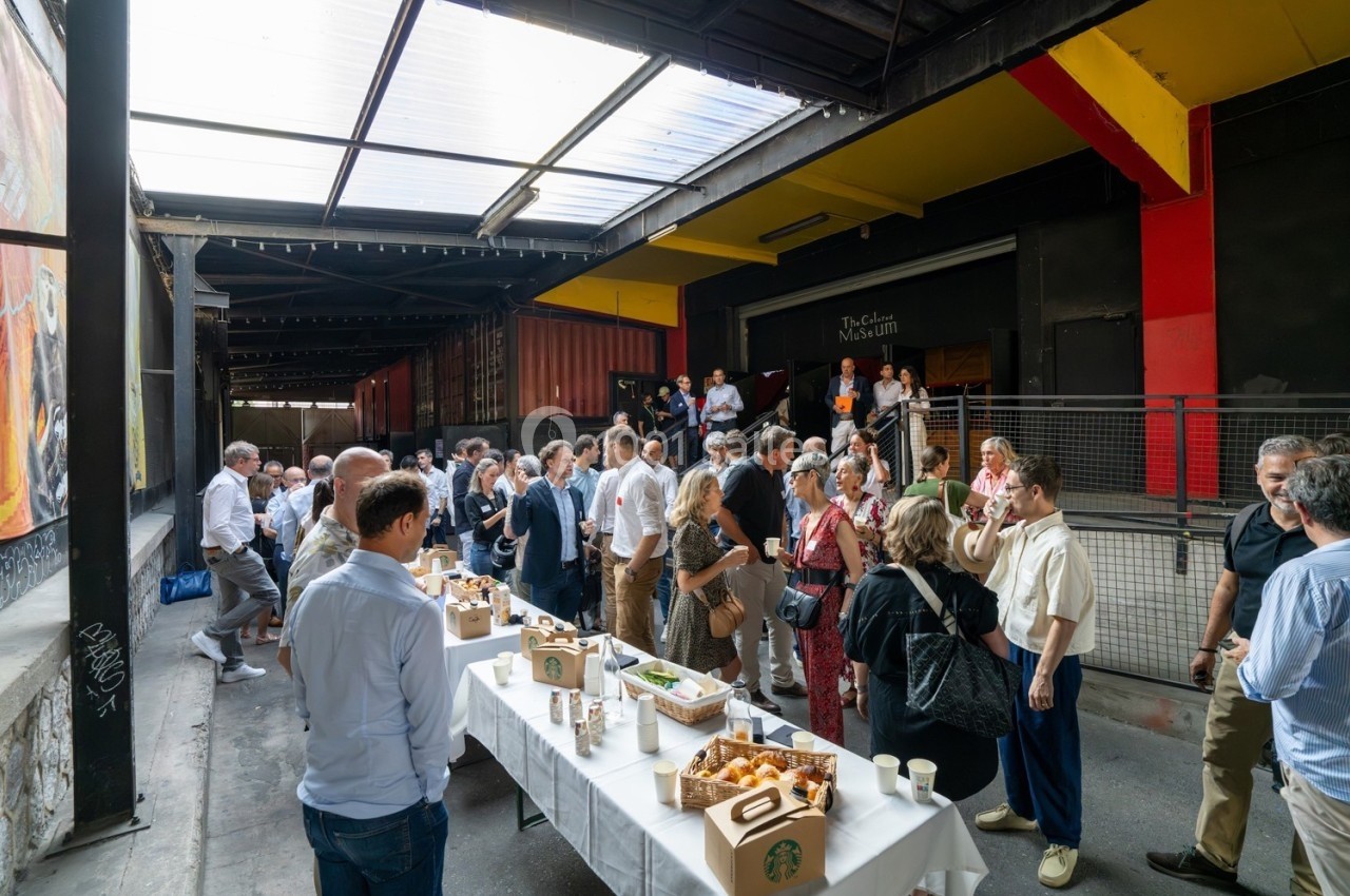 Groupe de personnes rassemblées autour de tables avec nourriture et boissons dans un espace extérieur couvert.