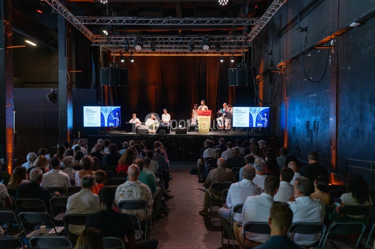 Conférence en intérieur avec plusieurs intervenants sur scène, face à un public assis dans une salle sombre.
