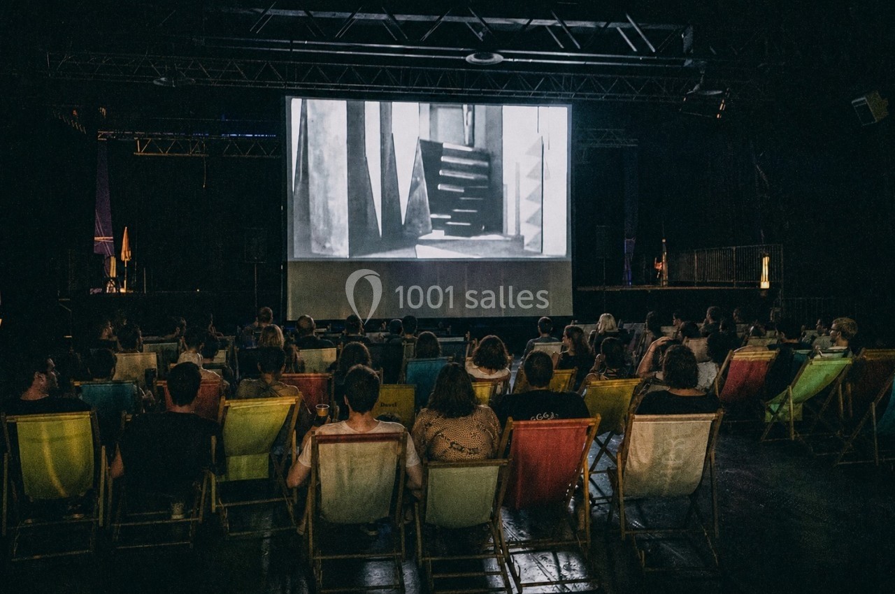 Des spectateurs assis sur des chaises longues regardent un film en noir et blanc projeté dans une salle sombre.