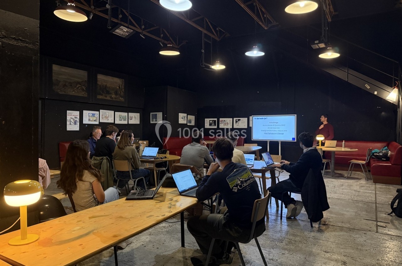 Des personnes assistent à une présentation dans une salle sombre, avec des ordinateurs portables sur des tables en bois.