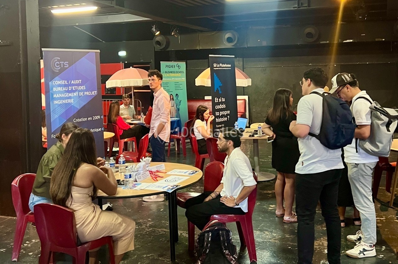 Des personnes échangent autour de stands d'information dans une salle avec des affiches et des tables.