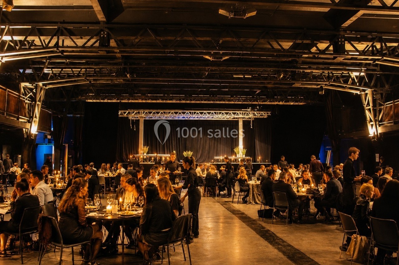 Salle industrielle avec des tables éclairées par des bougies, occupées par des convives lors d'un événement en soirée.