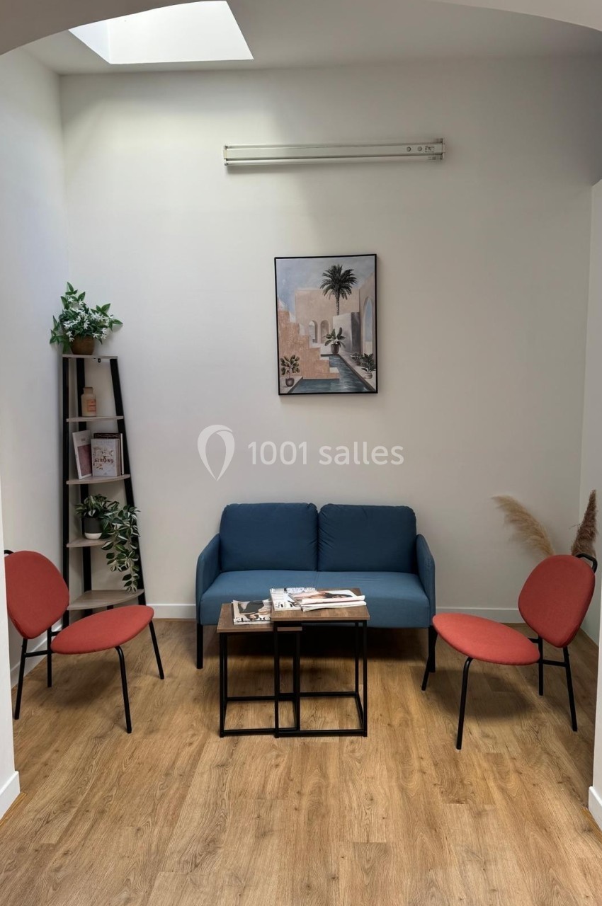 Espace d'attente avec canapé bleu, deux chaises rouges, table basse, étagère et décoration murale dans un intérieur lumineux.