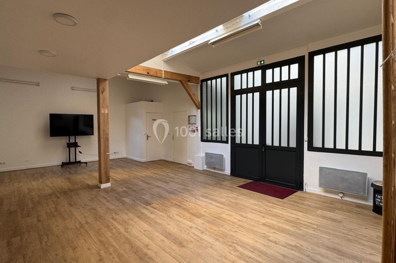 Salle lumineuse avec parquet en bois, grandes portes vitrées noires, écran sur pied et poutres apparentes.