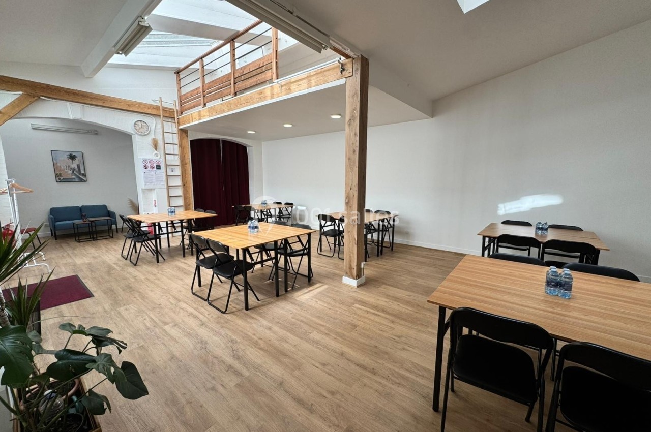 Salle lumineuse avec parquet, tables en bois, chaises noires, mezzanine en bois et plantes en décoration.