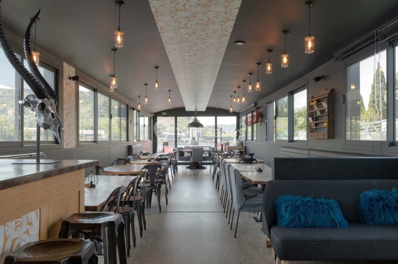 Salle de restaurant lumineuse avec grandes baies vitrées, mobilier en bois et métal, et décoration moderne.