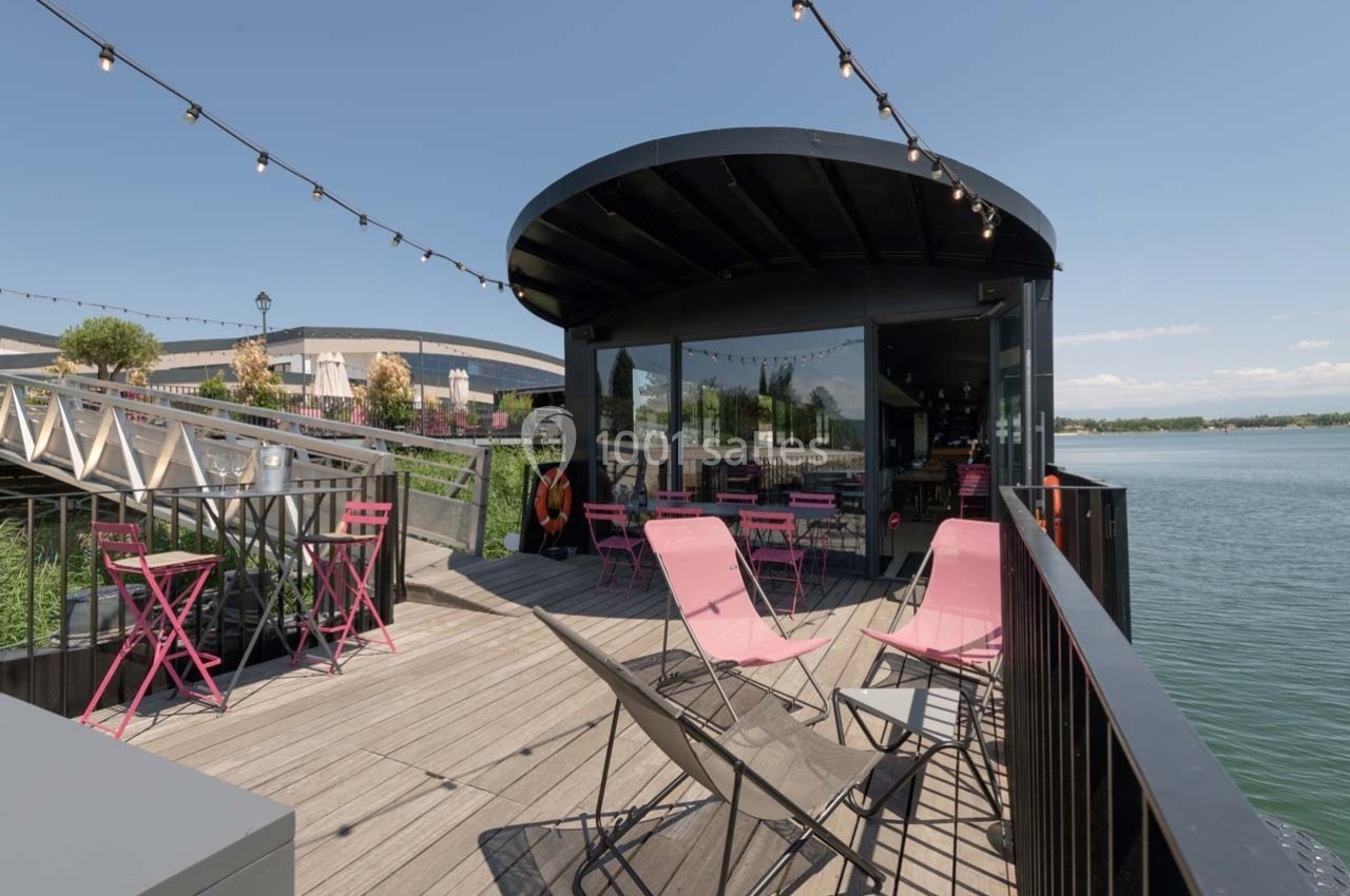 Terrasse en bois avec chaises roses, tables et guirlandes lumineuses, donnant sur un lac par une journée ensoleillée.