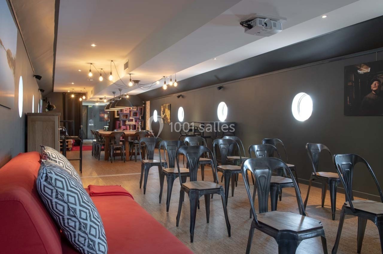 Salle avec des chaises en métal noir, un canapé rouge au premier plan et des luminaires suspendus.