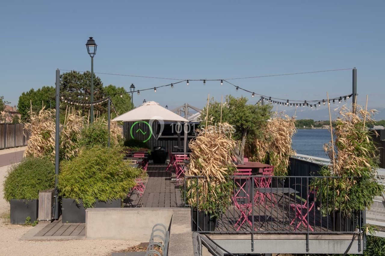 Terrasse en bois avec tables et chaises roses, entourée de plantes, surplombant un plan d'eau sous un ciel dégagé.