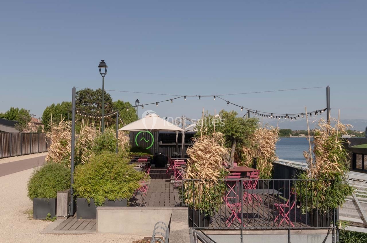 Terrasse extérieure avec tables et chaises roses, entourée de végétation, située au bord d'un plan d'eau.