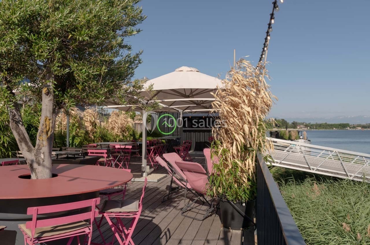 Terrasse en bois avec tables et chaises roses, parasol blanc, végétation et vue sur un plan d'eau.
