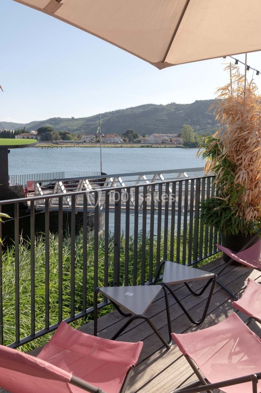 Terrasse avec chaises longues roses, vue sur un fleuve, des collines et un pont, partiellement ombragée par un parasol.