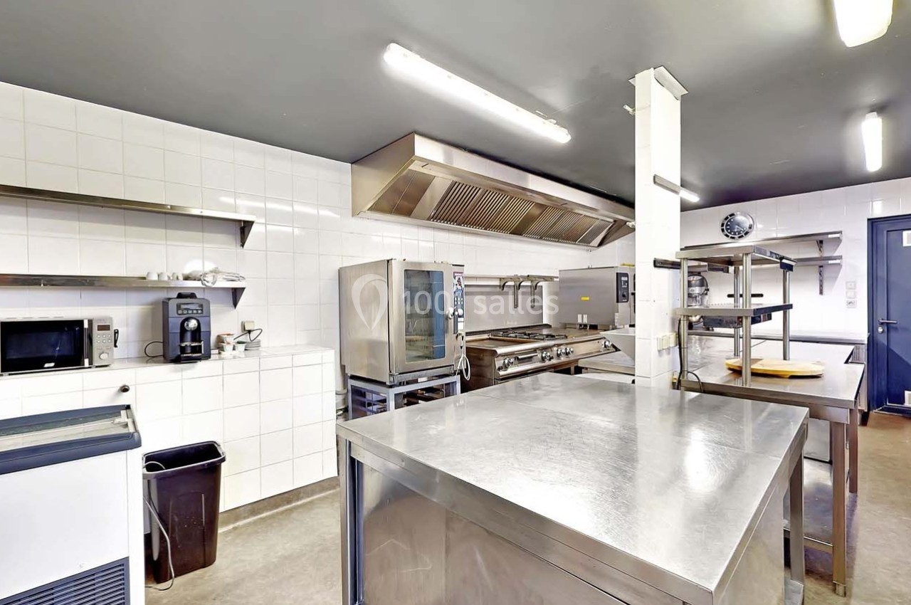 Cuisine professionnelle équipée avec plans de travail en inox, fours, étagères murales et éclairage fluorescent.