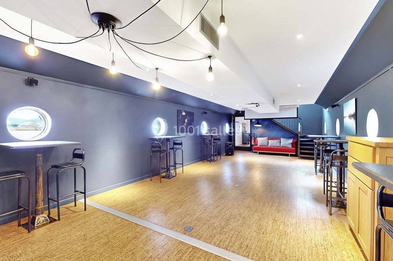 Salle lumineuse avec parquet, murs sombres, tables hautes, tabourets, hublots ronds et éclairage suspendu moderne.