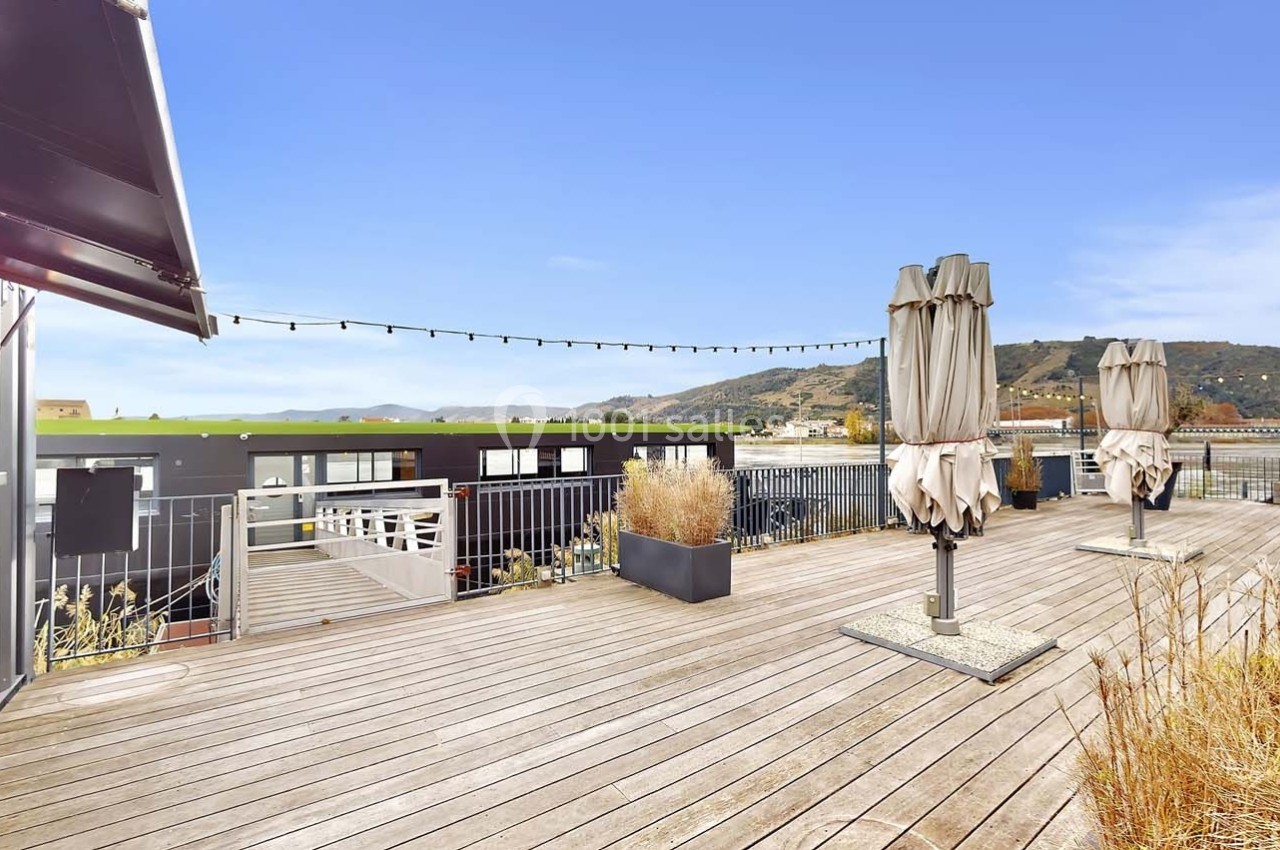 Terrasse en bois avec parasols fermés, plantes en pots et vue dégagée sur des collines sous un ciel bleu.