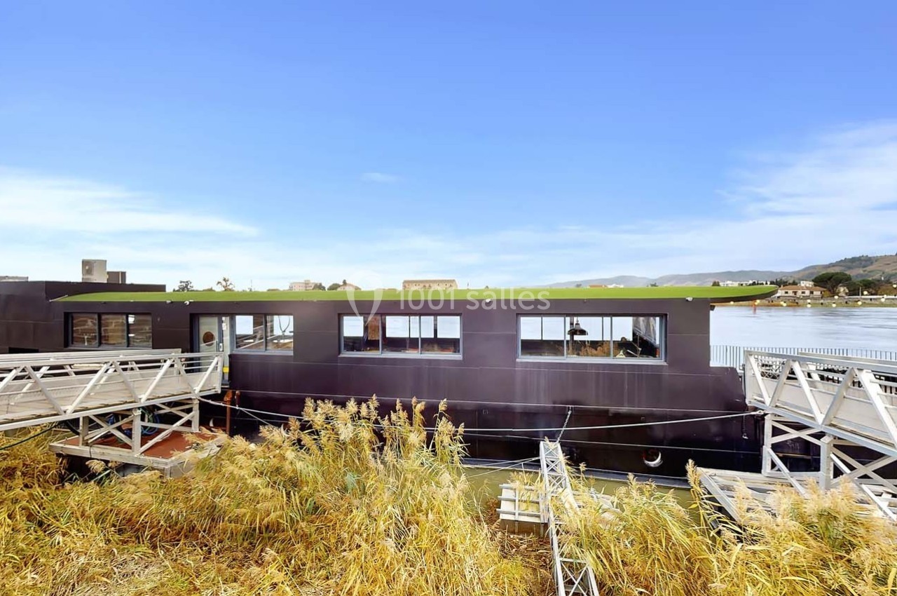 Bateau-maison moderne noir amarré près d'une passerelle, entouré de végétation et avec vue sur un plan d'eau.
