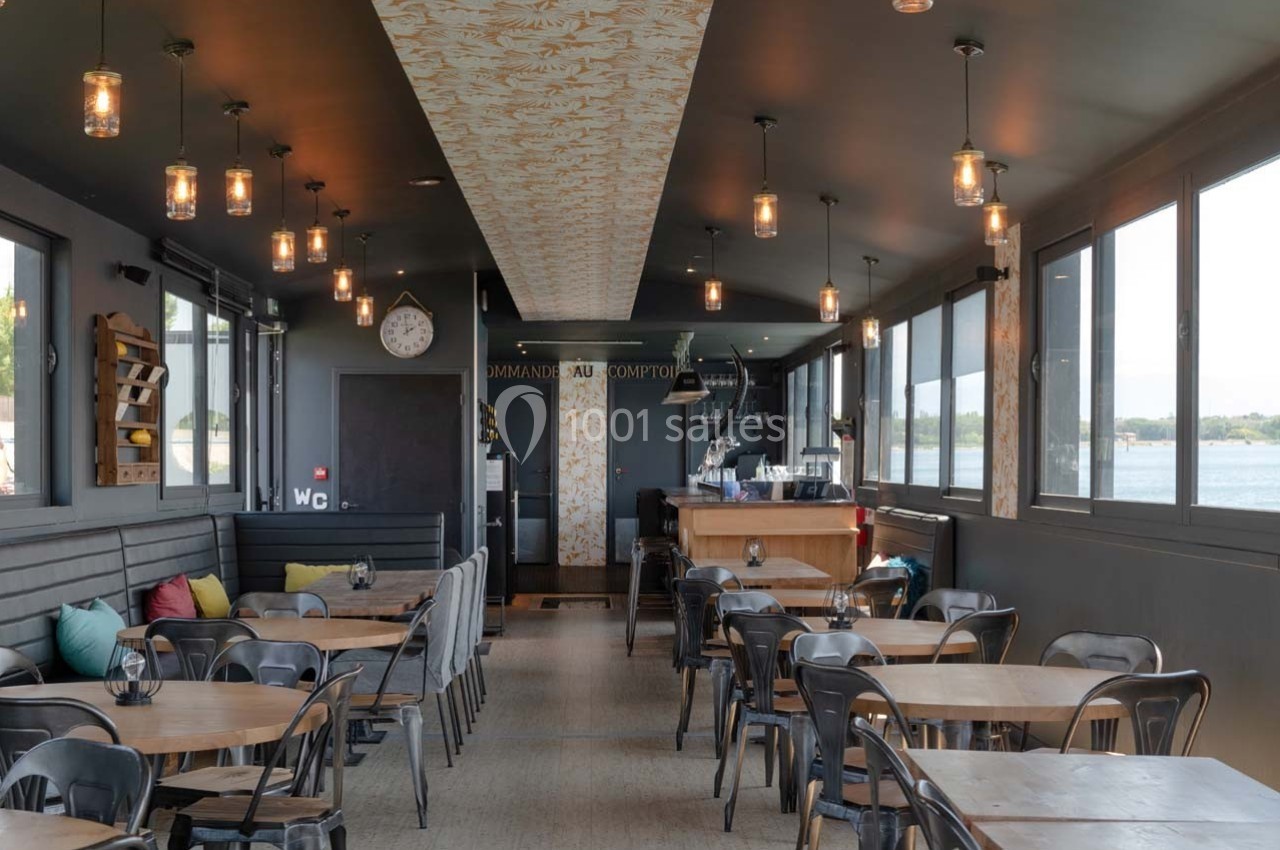 Salle de restaurant lumineuse avec tables en bois, chaises noires, banquettes et grandes fenêtres donnant sur l'extérieur.