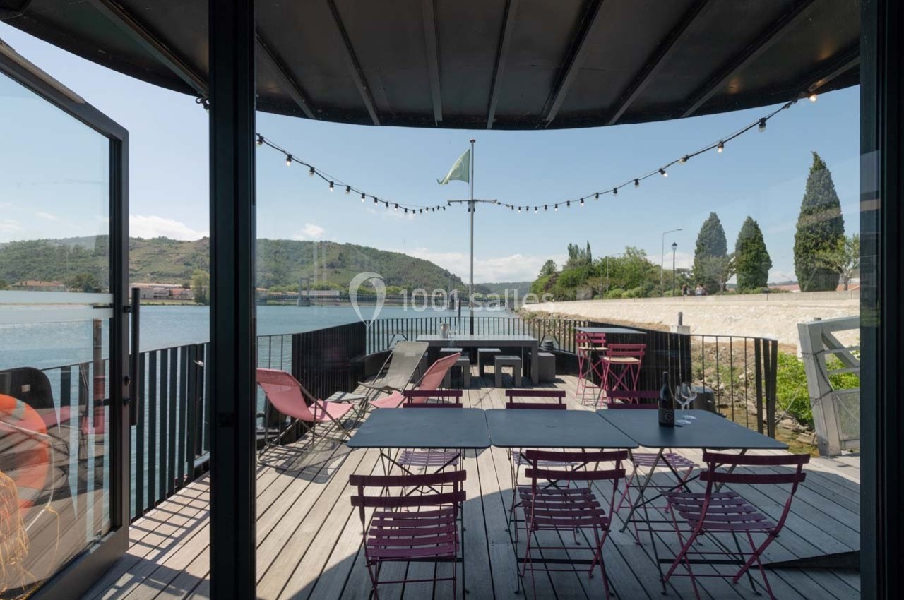 Terrasse en bois avec tables et chaises colorées, vue sur une rivière, collines et ciel dégagé en arrière-plan.