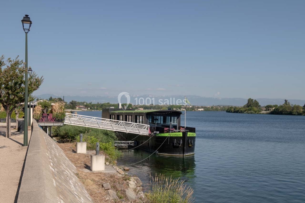 Une péniche amarrée le long d'une berge arborée, avec un paysage de rivière et des collines en arrière-plan.
