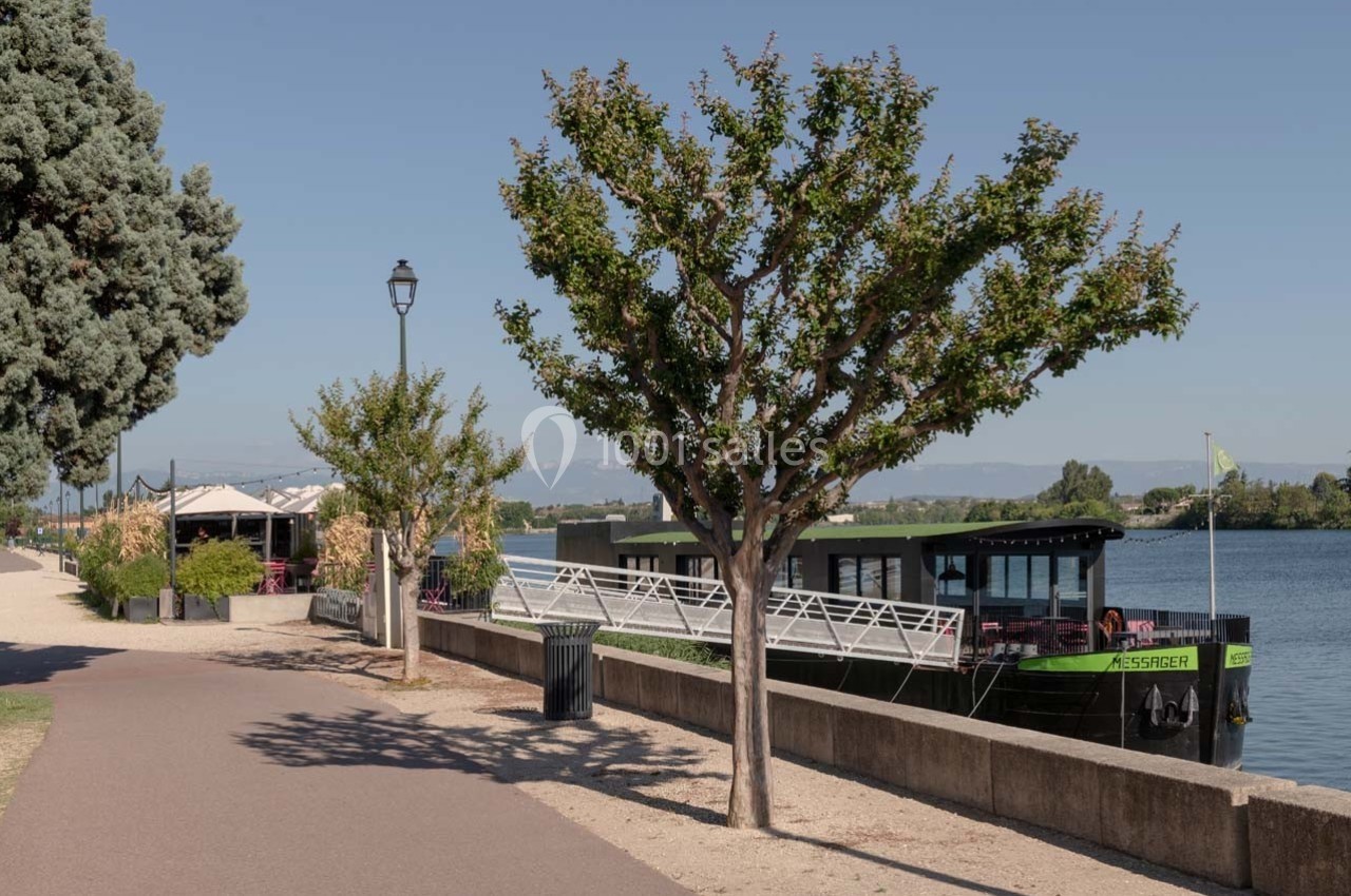 Allée bordée d'arbres longeant un fleuve avec une péniche amarrée et une terrasse de café à gauche.