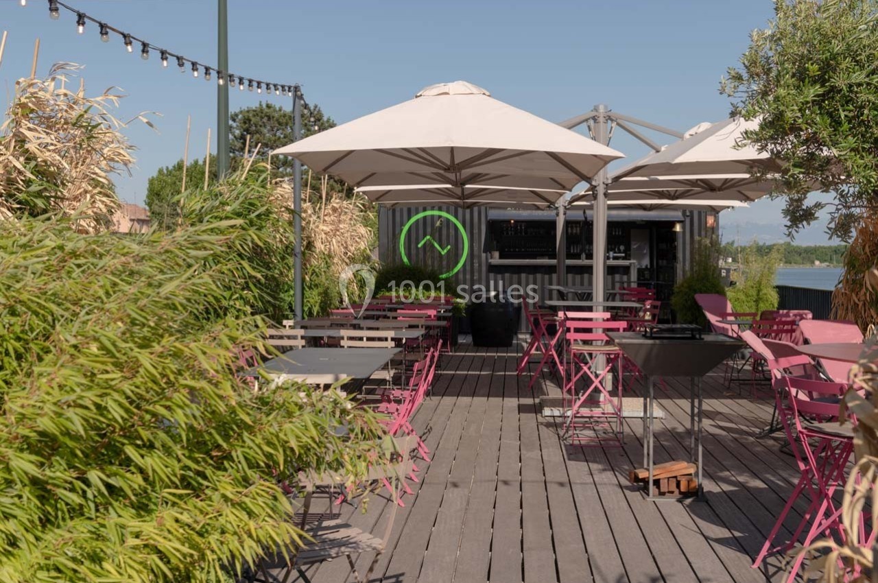Terrasse en bois avec tables et chaises roses, ombragée par des parasols blancs, entourée de végétation.