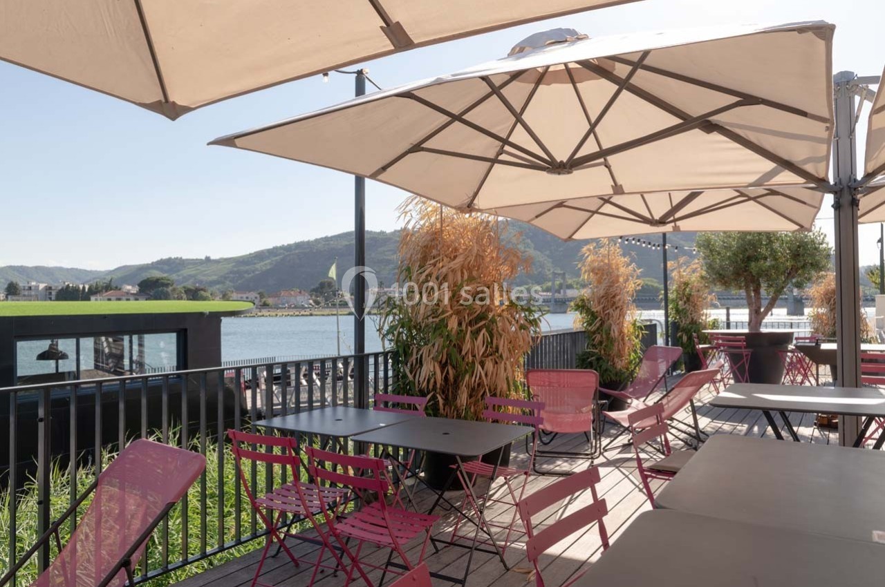Terrasse ensoleillée avec tables et chaises colorées, vue sur un paysage de colline et un plan d'eau.