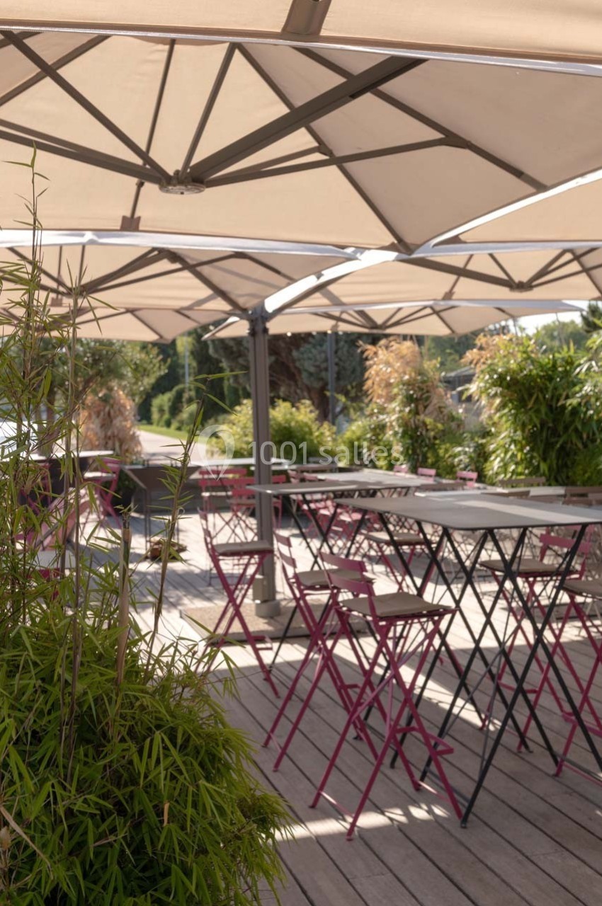 Terrasse ensoleillée avec tables et chaises pliantes roses sous de grands parasols, entourée de plantes vertes.