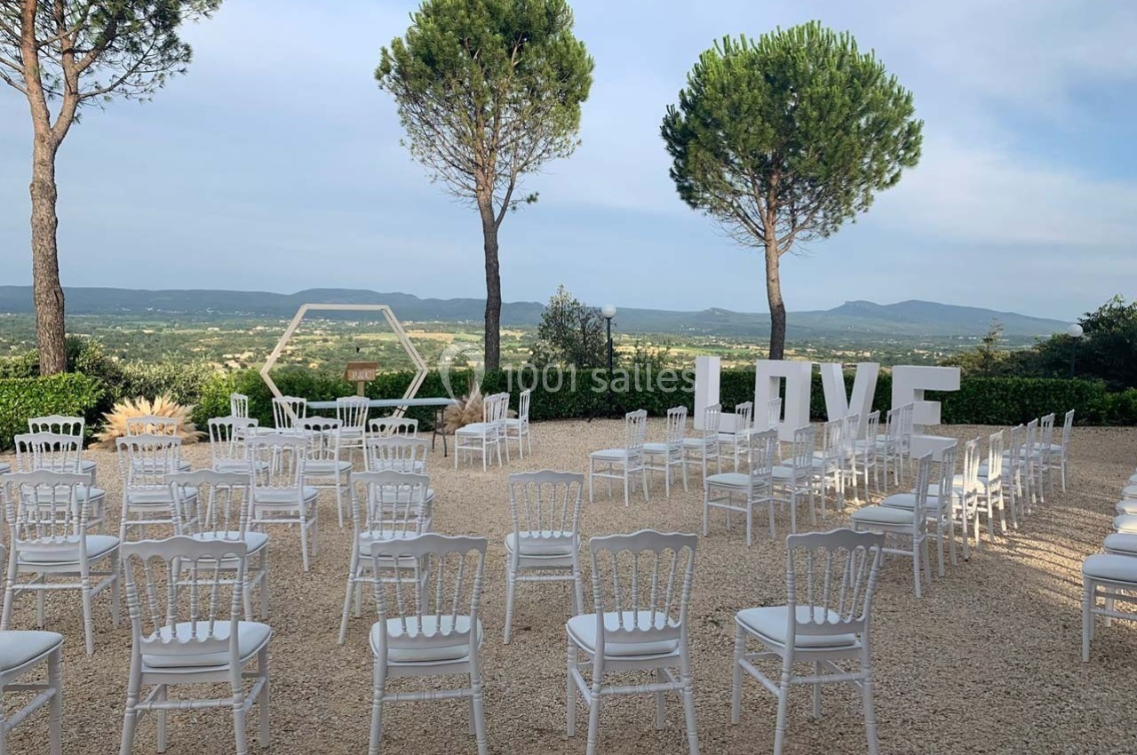 Chaises blanches disposées en extérieur pour une cérémonie, avec vue sur la campagne et décorations ’LOVE’.