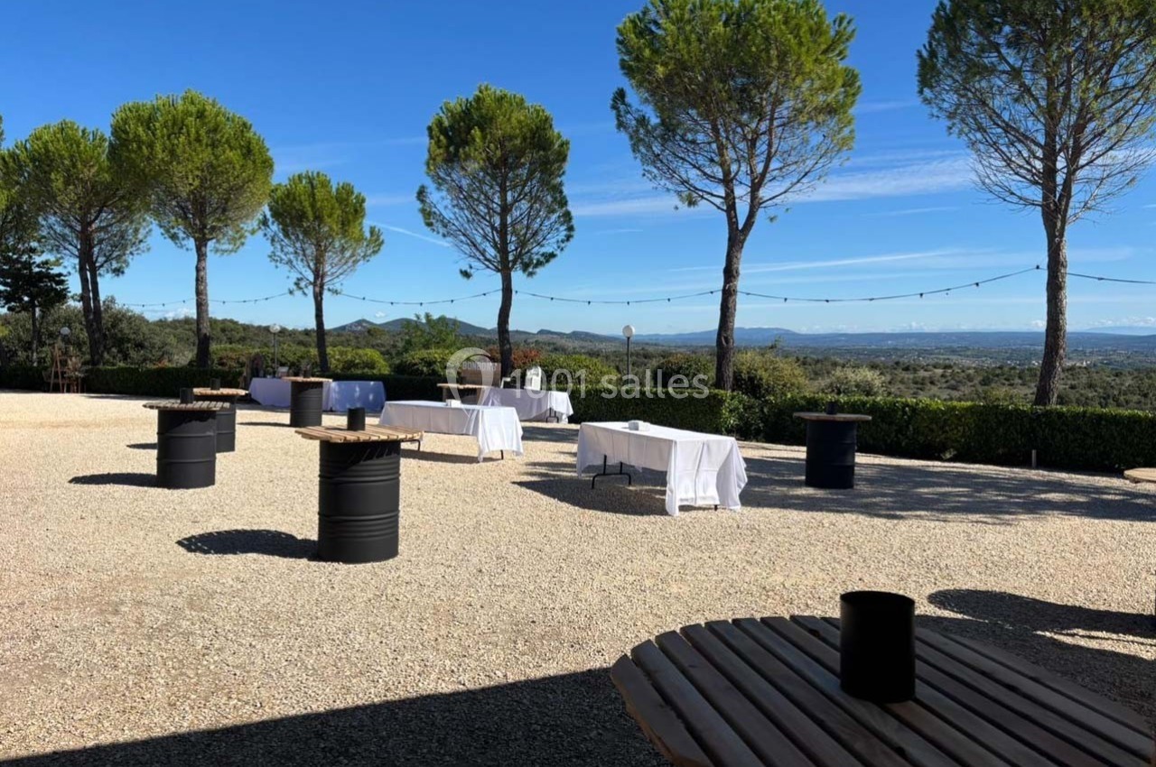 Terrasse extérieure avec tables en bois et tonneaux, vue sur un paysage vallonné et des arbres sous un ciel dégagé.