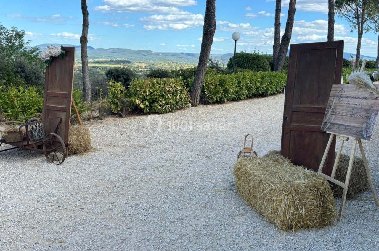 Chemin gravillonné bordé de végétation, décoré de bottes de paille, portes en bois et panneau d'accueil champêtre.