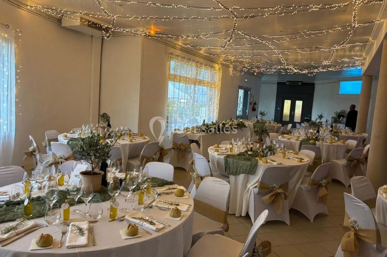 Salle de réception décorée pour un événement, avec tables rondes dressées, nappes blanches et guirlandes lumineuses au…