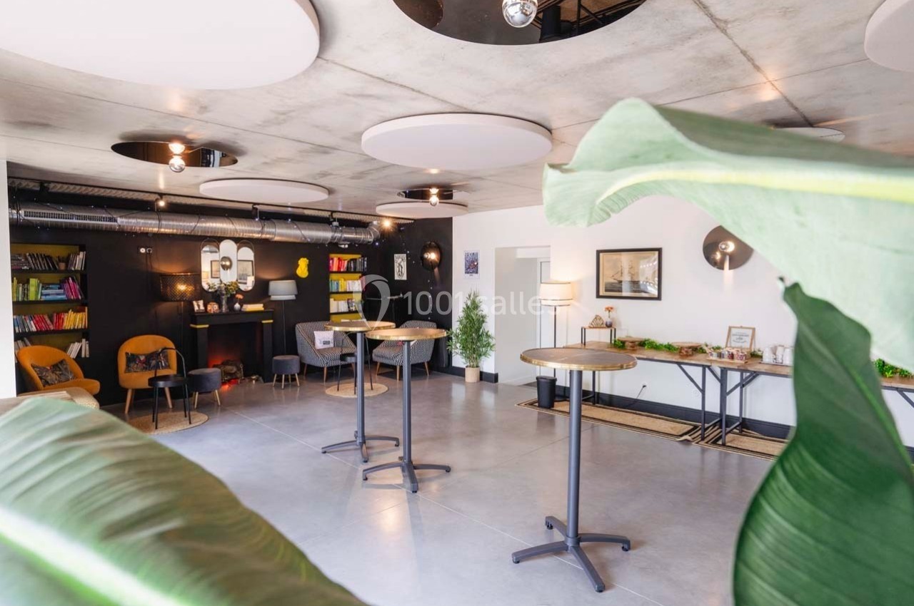 Espace intérieur lumineux avec tables hautes, fauteuils, étagères colorées et décorations végétales.