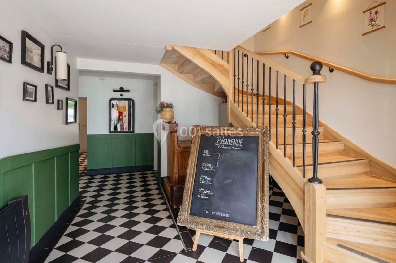 Entrée d'un bâtiment avec un escalier en bois, un tableau noir, sol en damier et décorations murales.