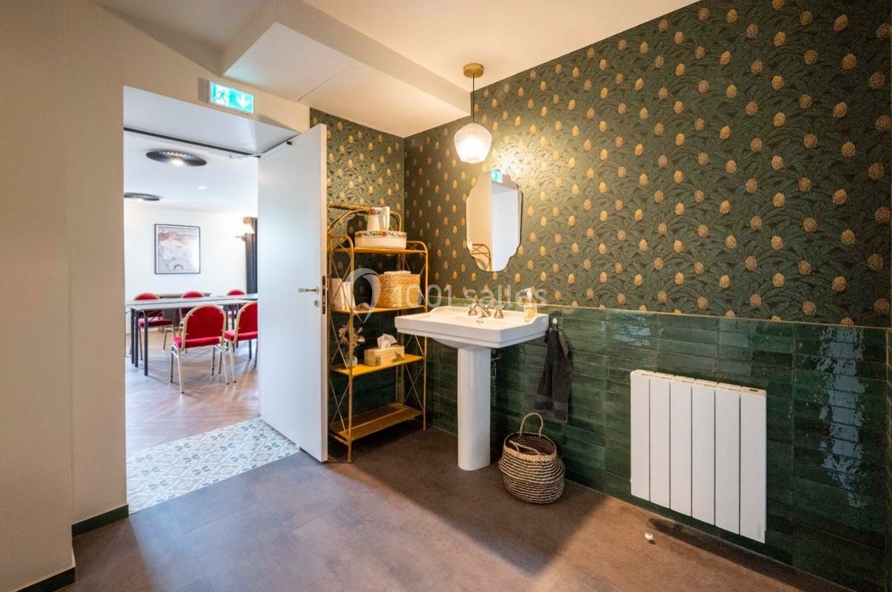 Salle de bain avec lavabo, miroir, étagère en osier, mur vert à motifs et vue sur une salle de réunion adjacente.