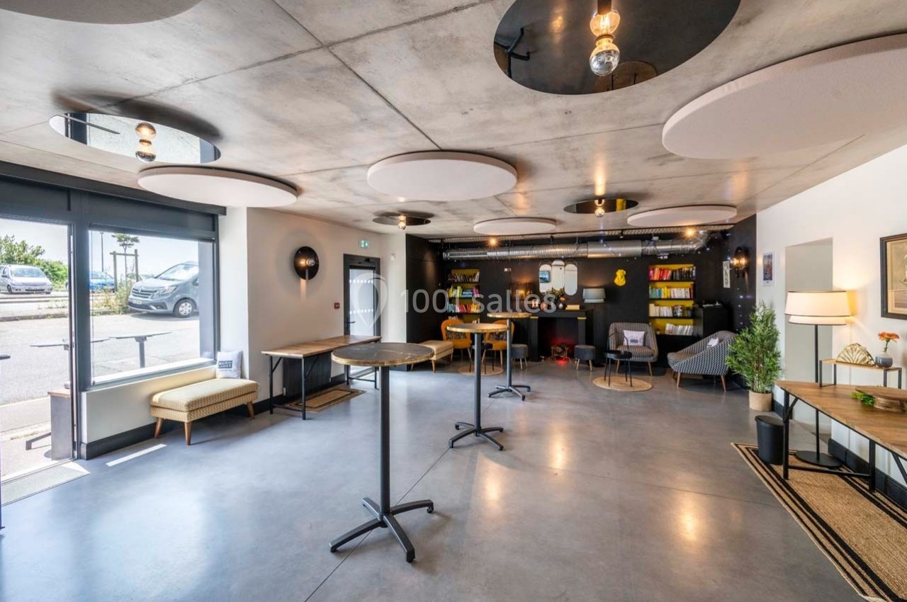 Salle lumineuse avec mobilier moderne, tables hautes, étagères colorées et grandes fenêtres donnant sur l'extérieur.
