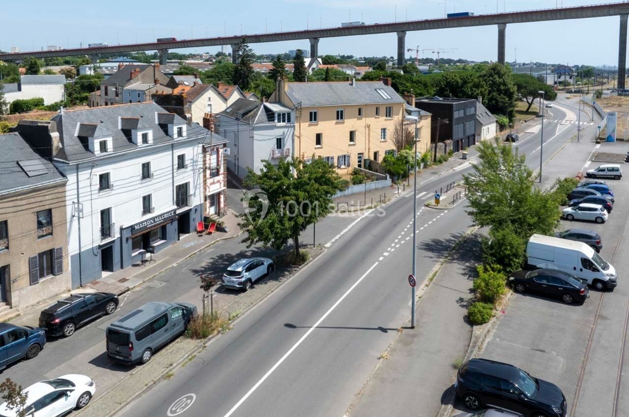 Rue bordée de maisons et de voitures stationnées, avec un pont surélevé en arrière-plan sous un ciel dégagé.