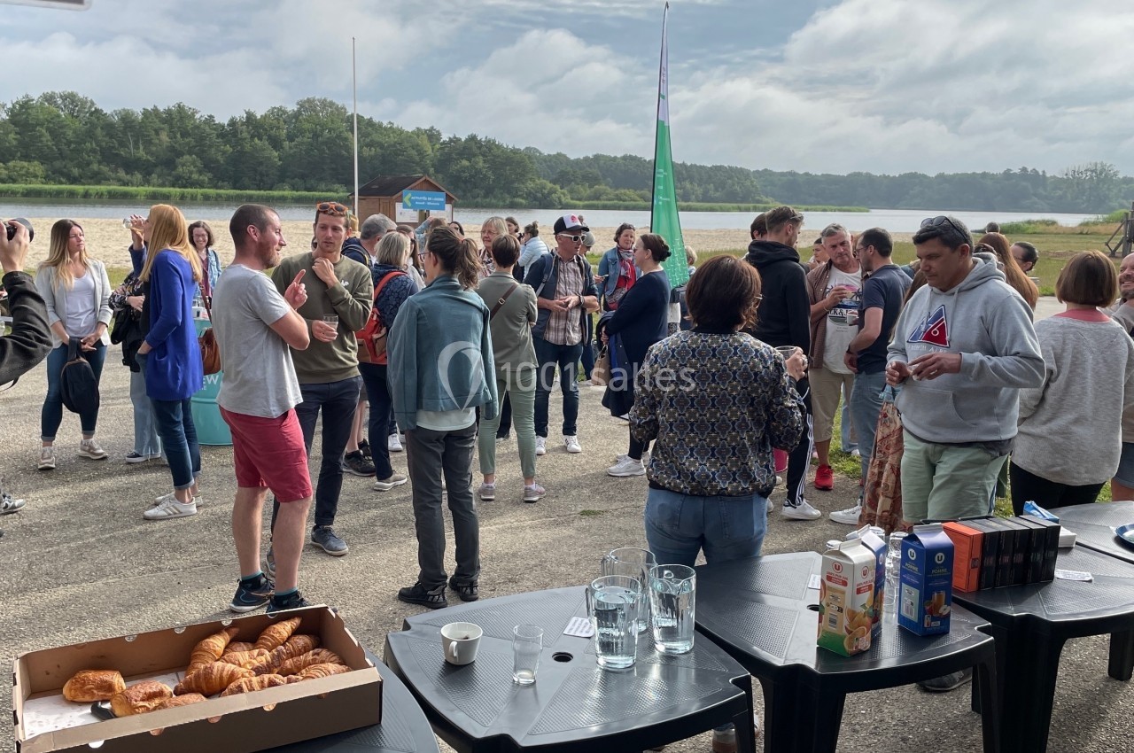 Groupe de personnes discutant en plein air près d'un lac, avec des tables contenant des boissons et des viennoiseries.