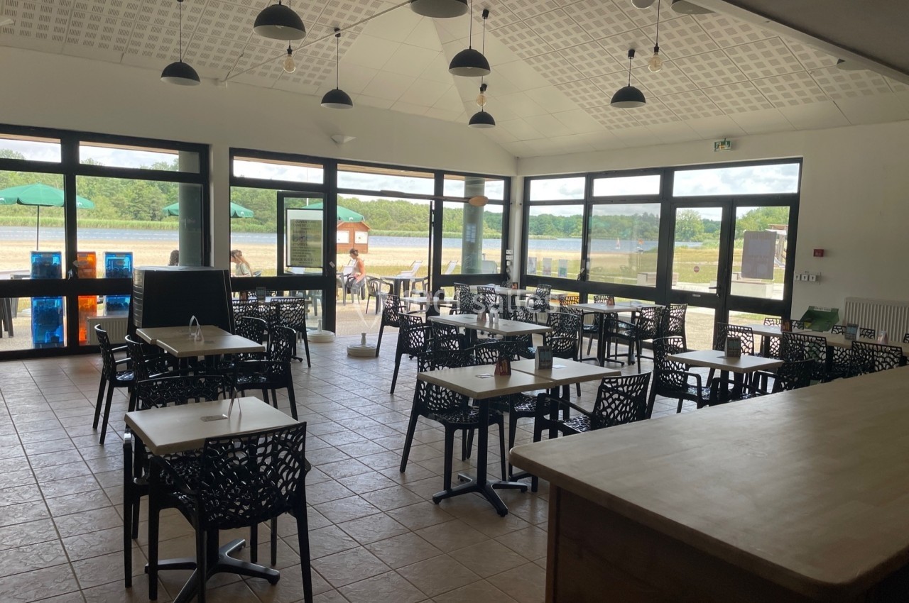 Salle lumineuse avec tables et chaises noires, grandes baies vitrées donnant sur un espace extérieur verdoyant.