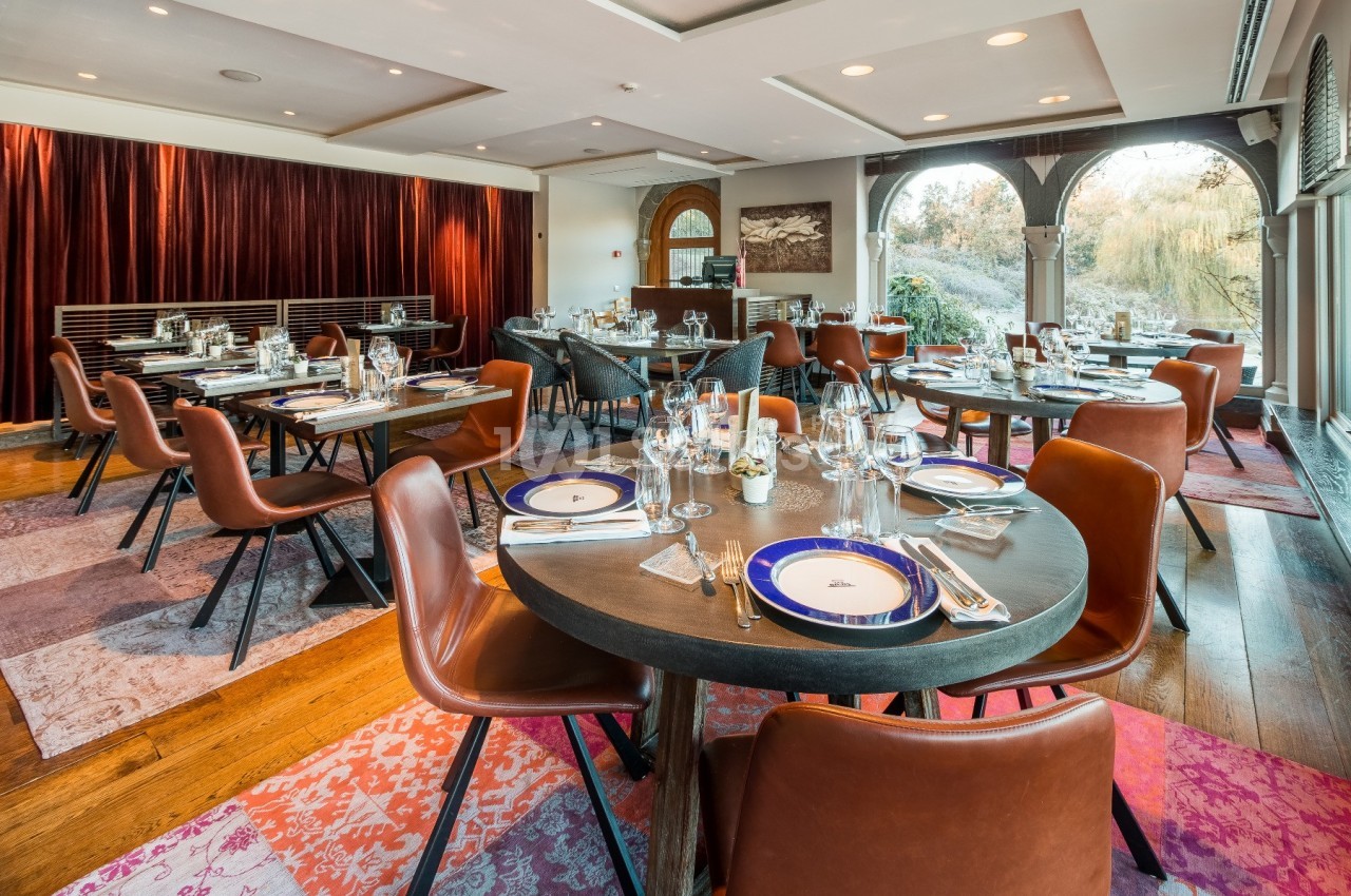 Salle de restaurant élégante avec tables dressées, chaises en cuir marron et grandes baies vitrées donnant sur un jardin.