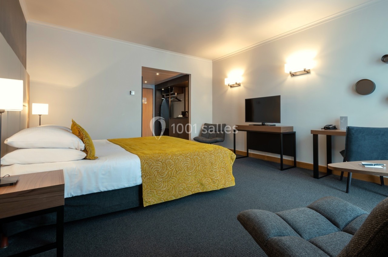 Chambre d'hôtel moderne avec lit double, bureau, télévision, fauteuils et éclairage chaleureux.