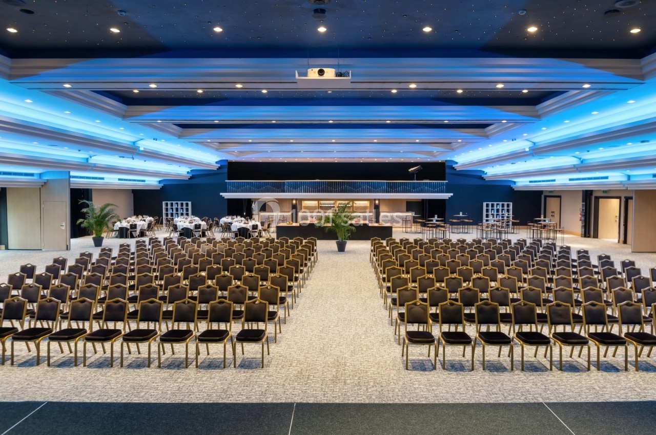 Salle de conférence spacieuse avec rangées de chaises alignées face à une scène et un espace central dégagé.