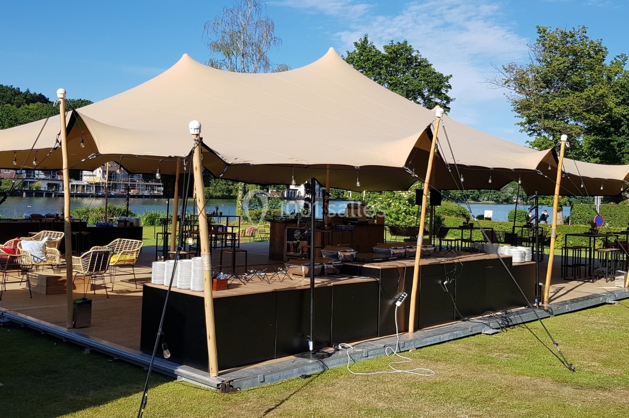 Tente beige montée sur une structure en bois, aménagée avec des tables et chaises, située dans un espace vert près d'un lac.