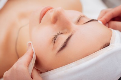 Femme allongée recevant un soin du visage avec un bandeau blanc, une personne nettoie sa peau avec des cotons.