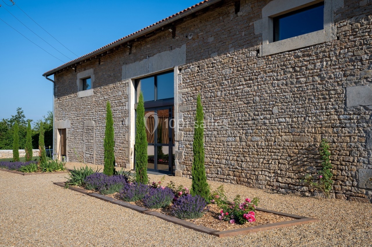 Façade en pierre d'un bâtiment rénové avec de grandes fenêtres, bordée de parterres fleuris et de cyprès.